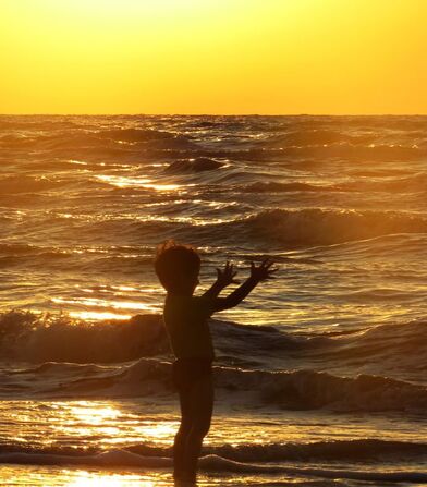 Child on beach shoreline with hands in air and sun setting in background. Being outdoors helps children and teens cope with anxiety and OCD