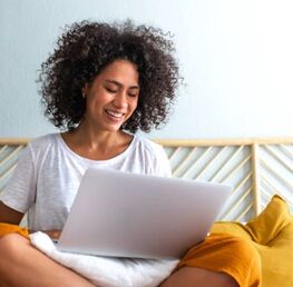 Mujer sonriente sentada arriba de su cama, mirando su laptop. Dra. Penela ofrece terapia online en español para adultos en la Florida que sufren de ansiedad o el TOC o OCD.