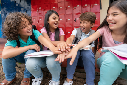 Four children sit on a curb and place their hands together while laughing and smiling at school.